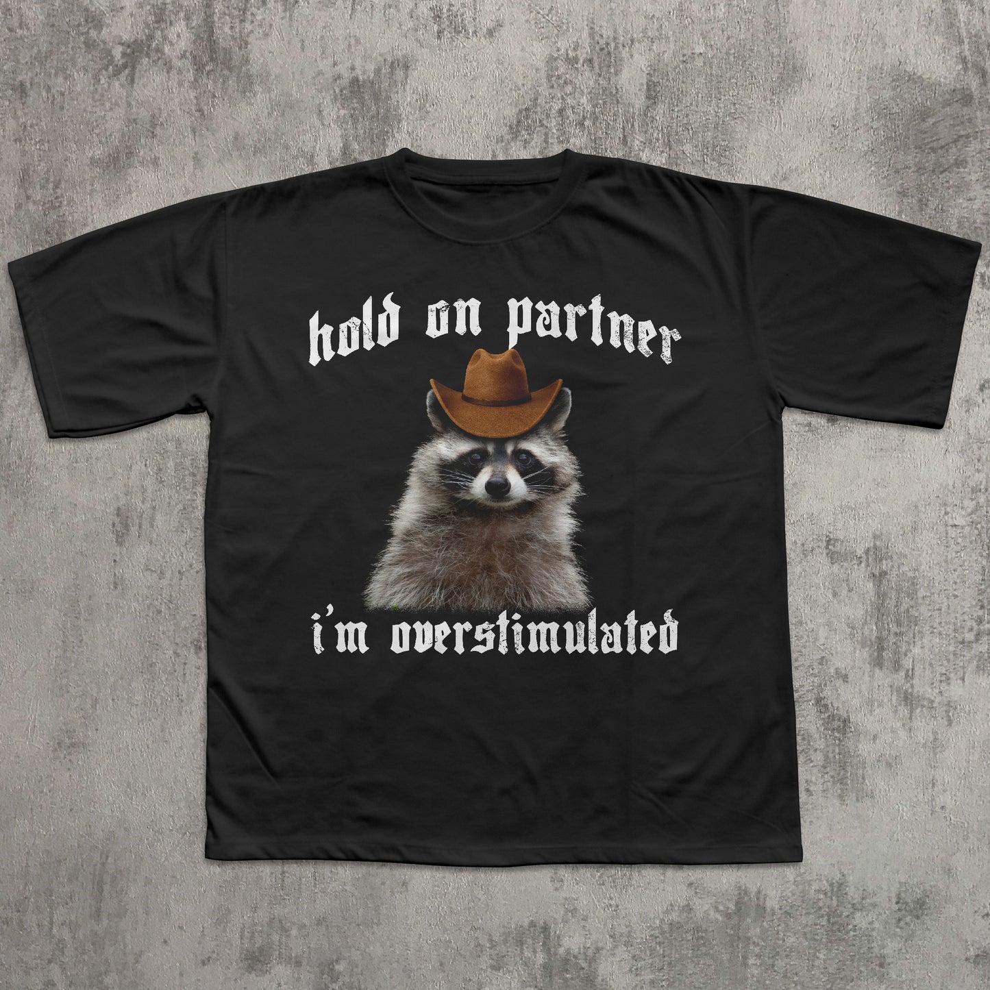 Black t-shirt with a raccoon wearing a cowboy hat and text on a textured gray background
