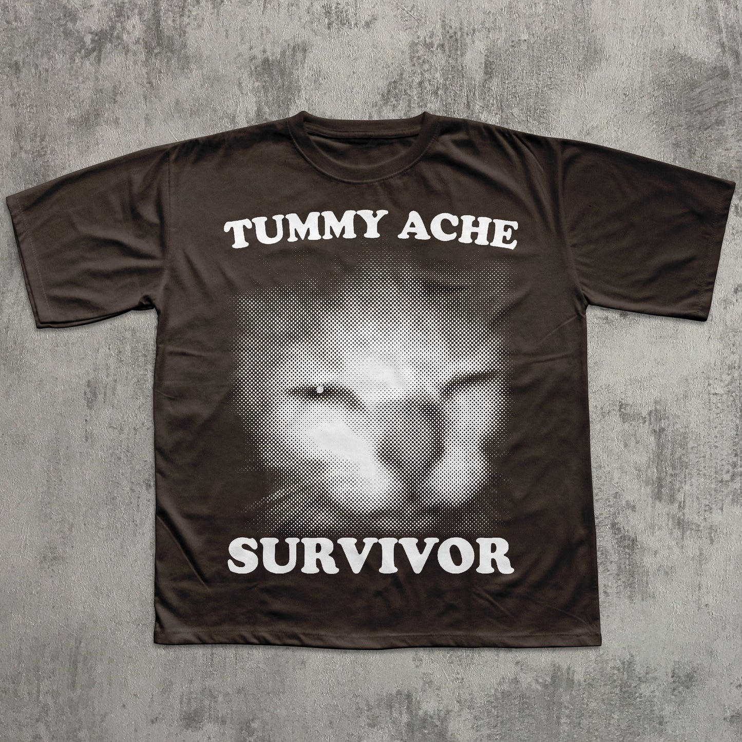 Brown t-shirt with a graphic of a cat's face and text 'TUMMY ACHE SURVIVOR' on a textured gray background