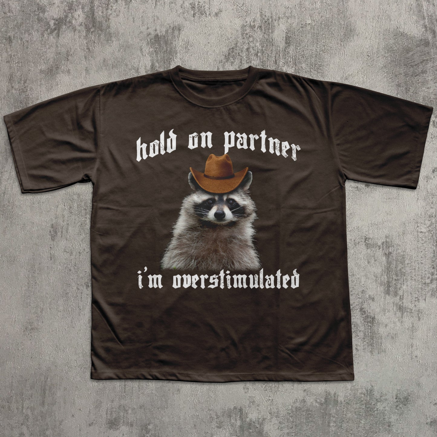 Brown t-shirt with a raccoon wearing a cowboy hat and text on a textured gray background