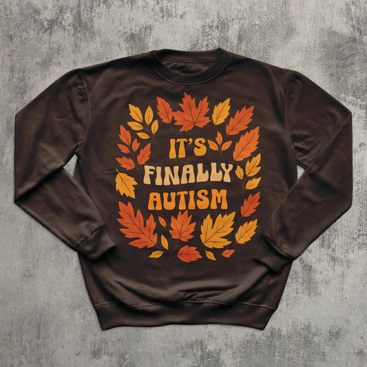 Brown sweatshirt with autumn leaf design and 'It's Finally Autism' text on a gray textured background