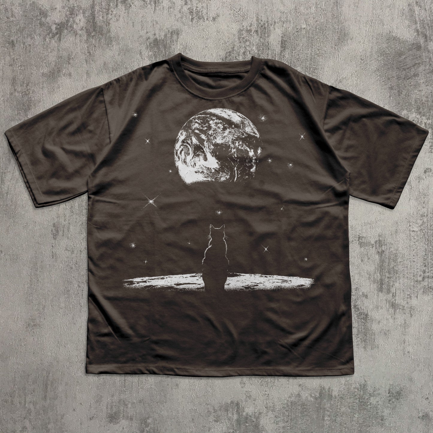 Brown t-shirt with a graphic of a cat on a surfboard and a moon in the background, laid flat on a textured gray surface.