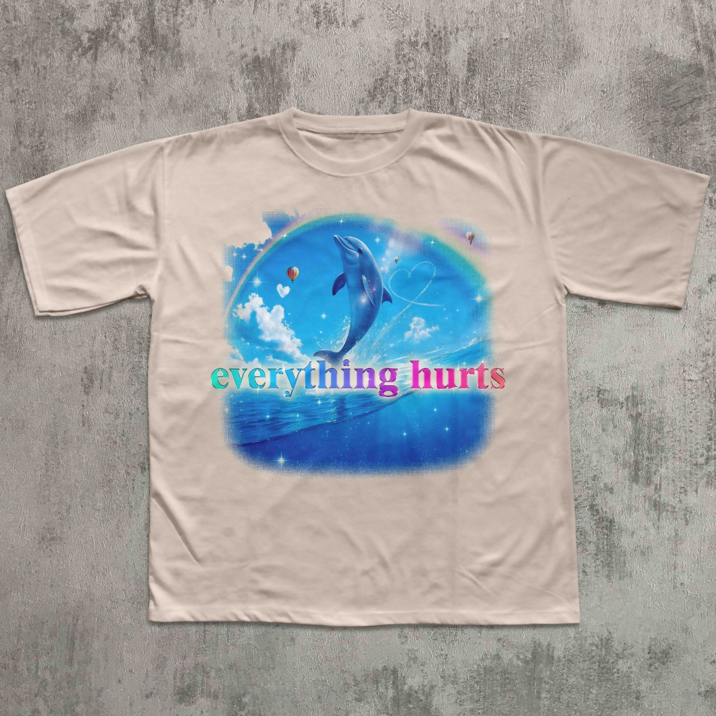 Beige t-shirt with a blue graphic of a dolphin and text 'everything hurts' on a textured gray background