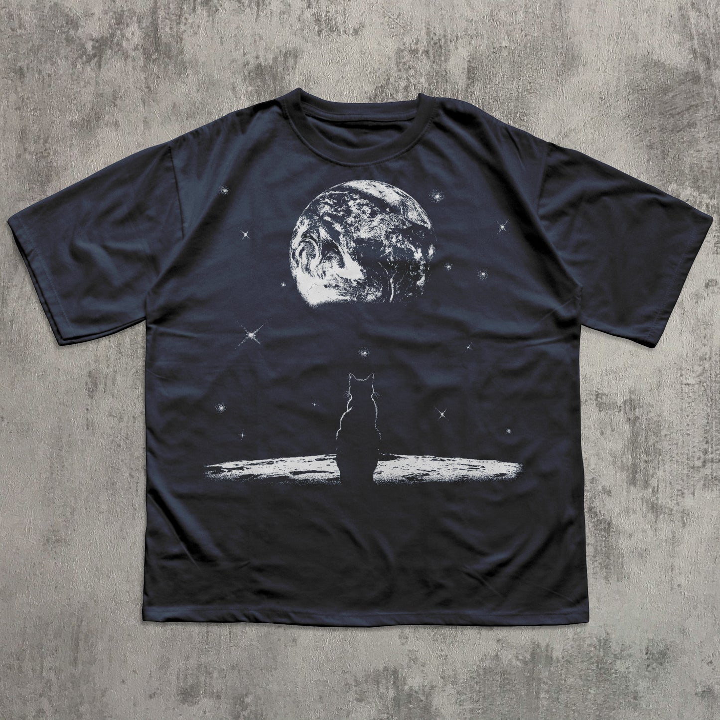 Black t-shirt with a graphic of a cat on a surfboard and Earth in the background, laid flat on a textured surface.