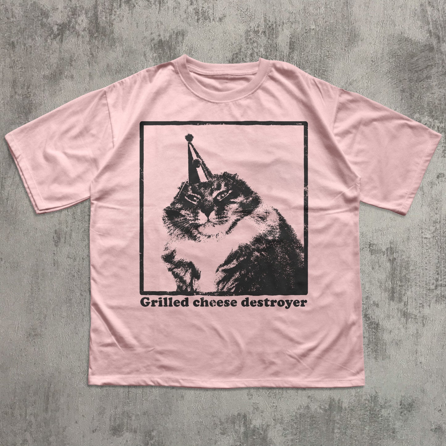 Pink t-shirt with a graphic of a cat and text 'Grilled cheese destroyer' on a textured gray background