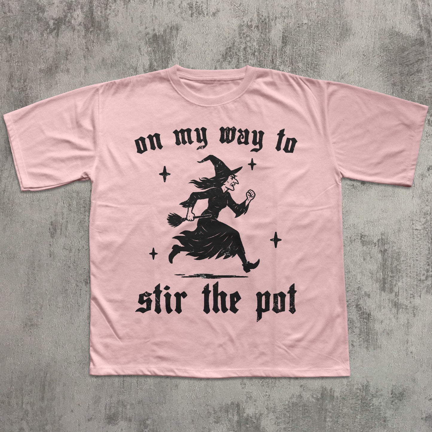 Pink t-shirt with a witch graphic and text on a gray concrete background