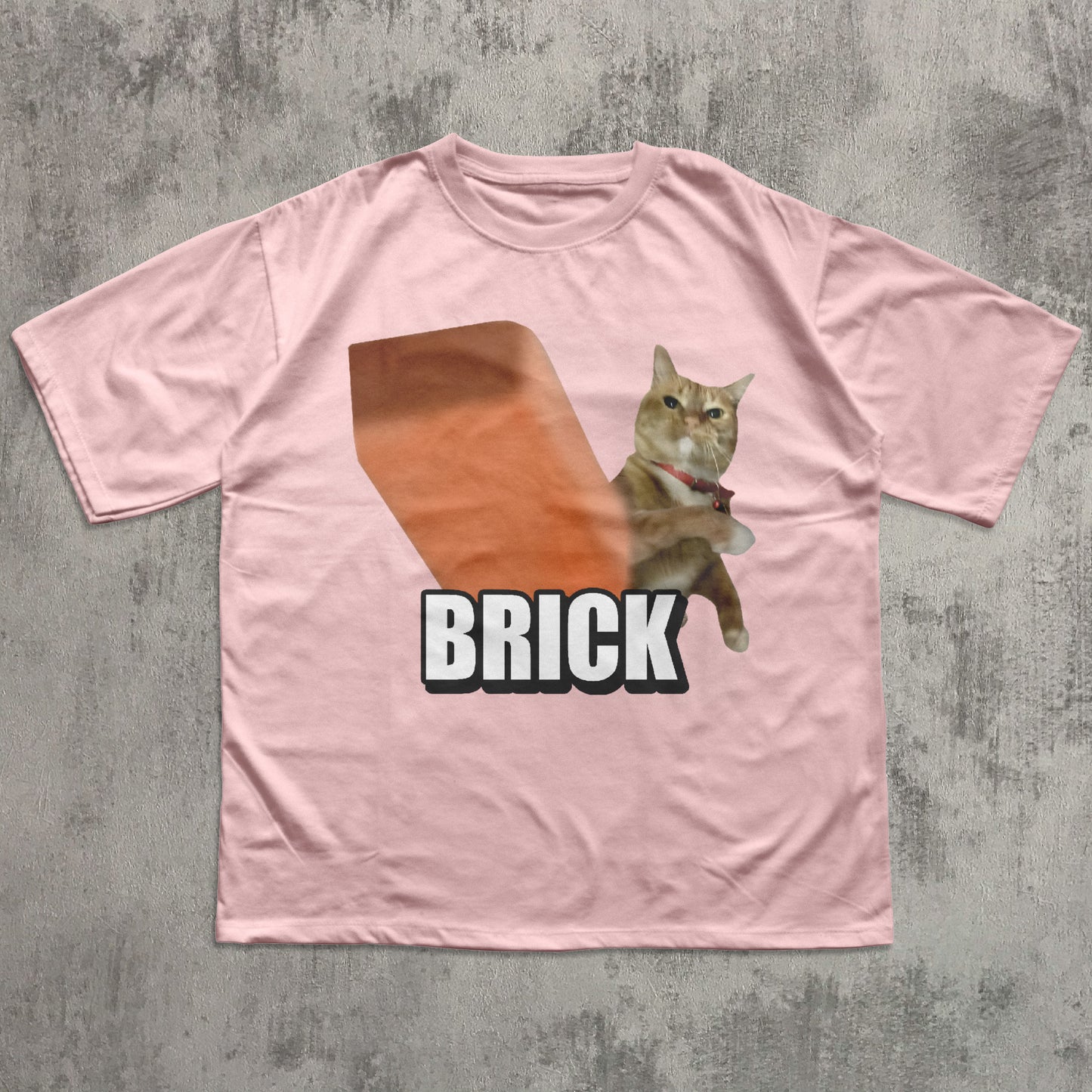 Pink t-shirt with a cat holding a brick graphic and 'BRICK' text on a textured gray background