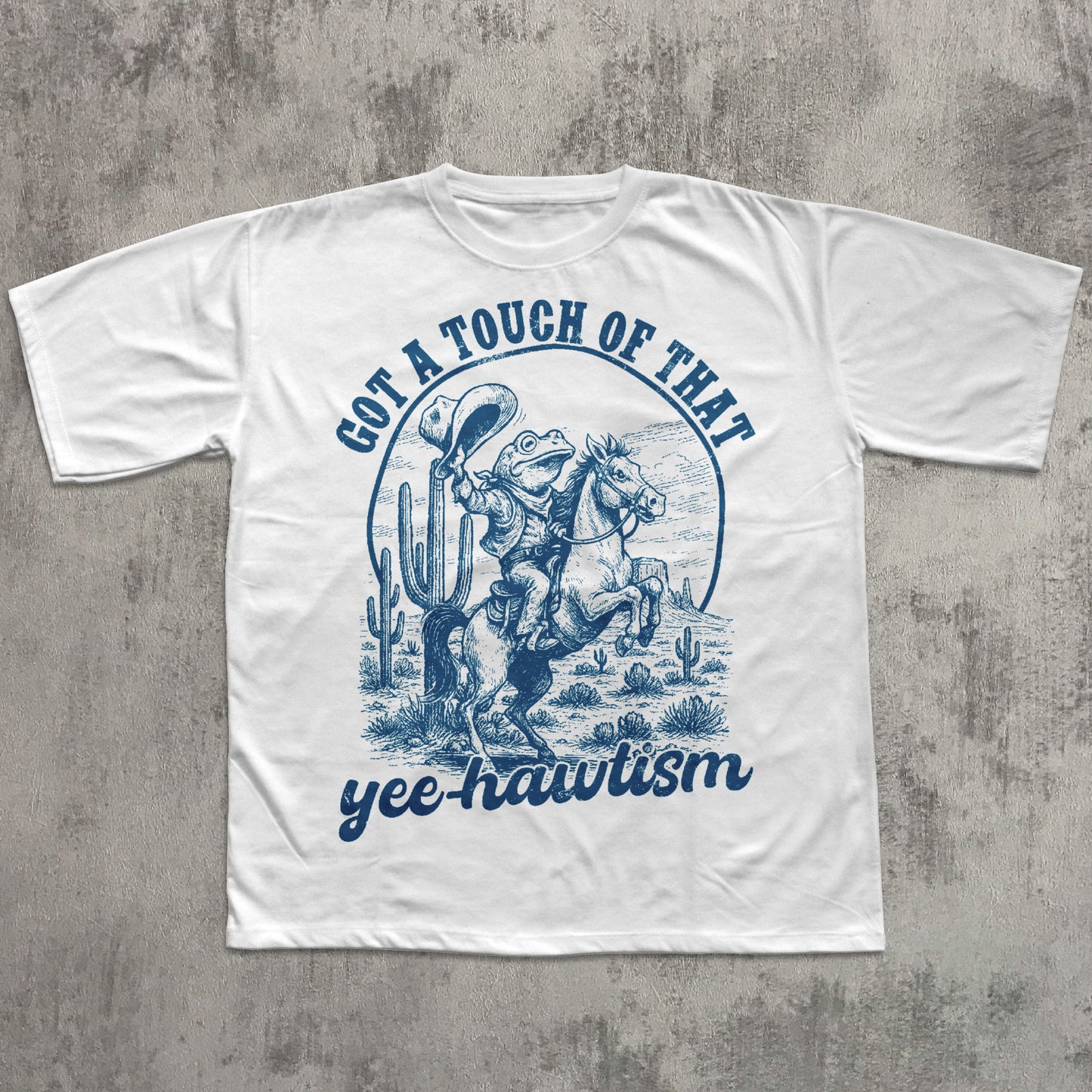 White T-shirt featuring a blue vintage-style illustration of a cowboy frog riding a rearing horse in a desert scene with cacti, with the text ‘Got a touch of that yee-hawtism’ above and below the graphic.
