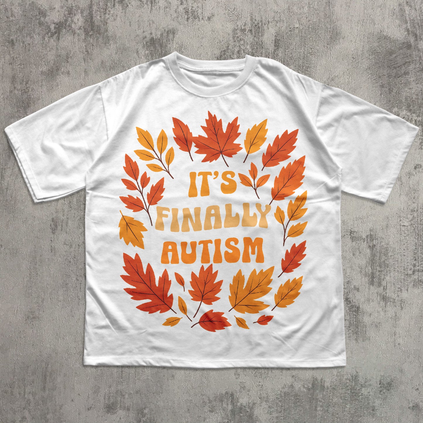 White t-shirt with autumn leaf design and 'It's Finally Autism' text on a concrete surface