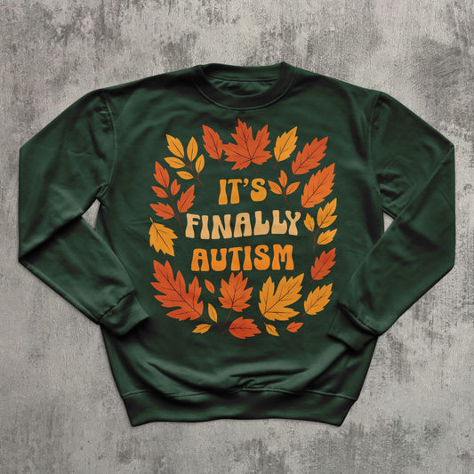 Green sweatshirt with autumn leaf design and 'It's Finally Autism' text on a gray textured background
