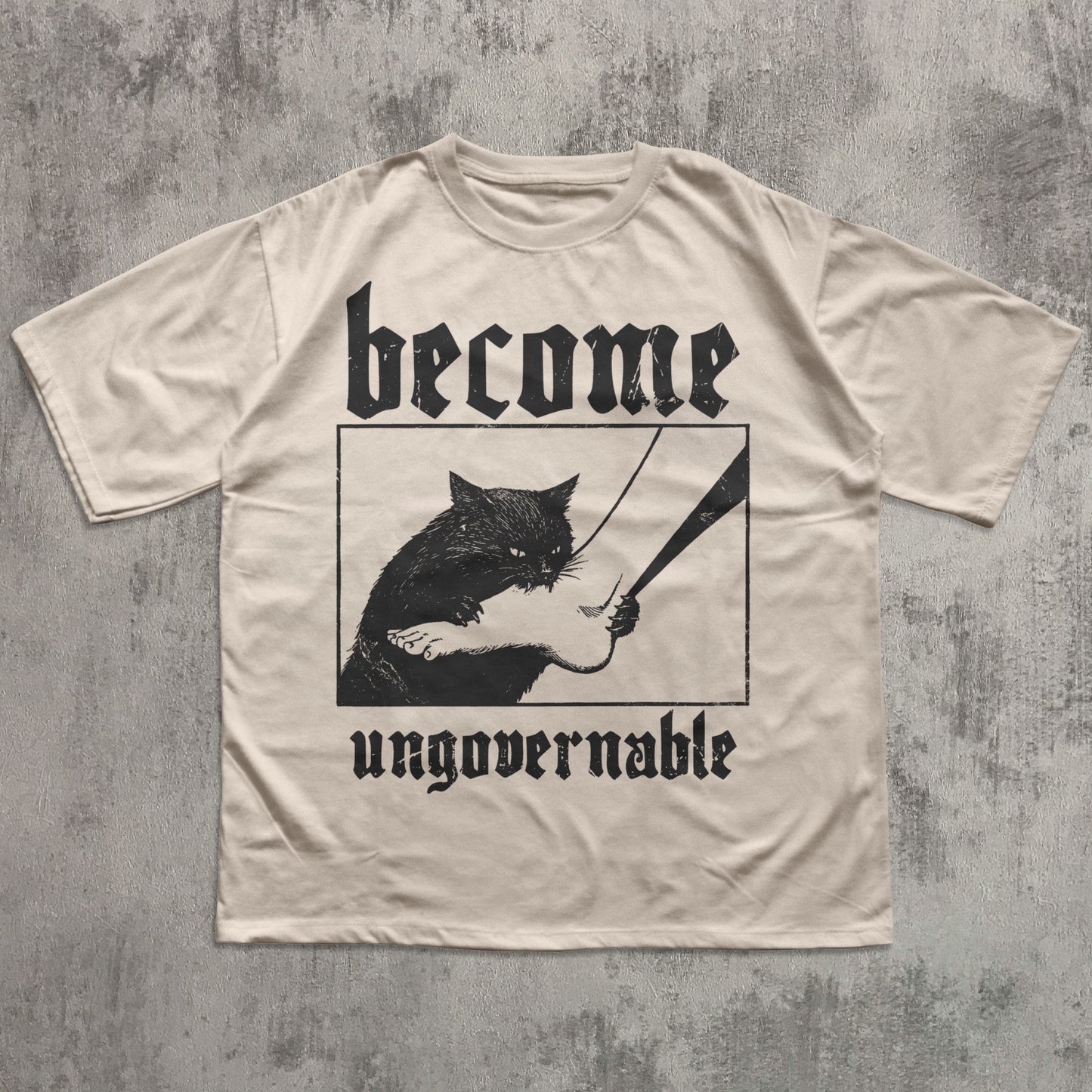 Beige t-shirt with black graphic of a cat holding a sword and text 'become ungovernable' on a textured gray background