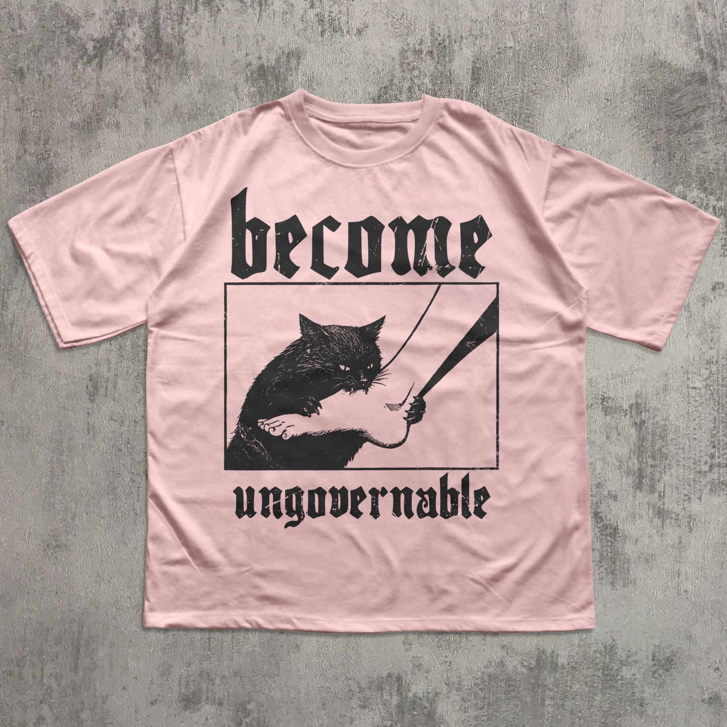 Pink t-shirt with a graphic of a cat holding a sword and text 'become ungovernable' on a concrete surface.