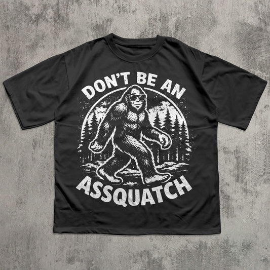 Black t-shirt with a graphic of a bigfoot and text 'Don't Be An Assquatch' on a concrete surface.