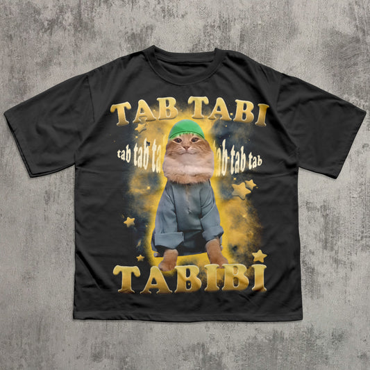 Funny black t-shirt with a graphic of a cat wearing a green hat and blue pants, surrounded by stars, on a concrete surface.