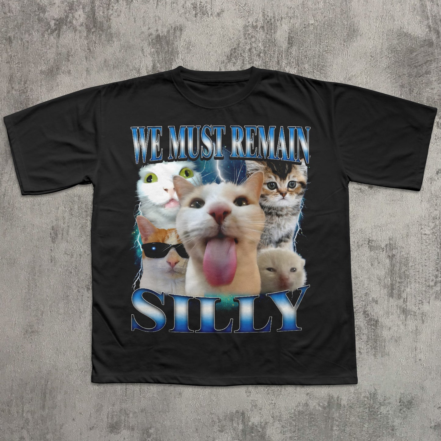 Black t-shirt with cartoon cats and text 'WE MUST REMAIN SILLY' on a textured gray background