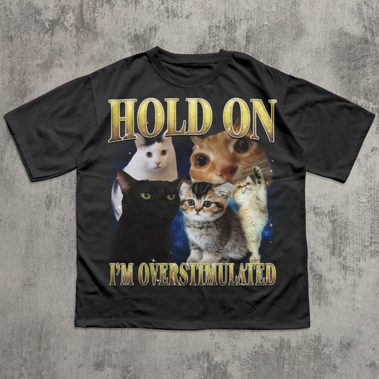 Black t-shirt with cat graphic and text 'Hold on I'm overstimulated' on a textured gray background