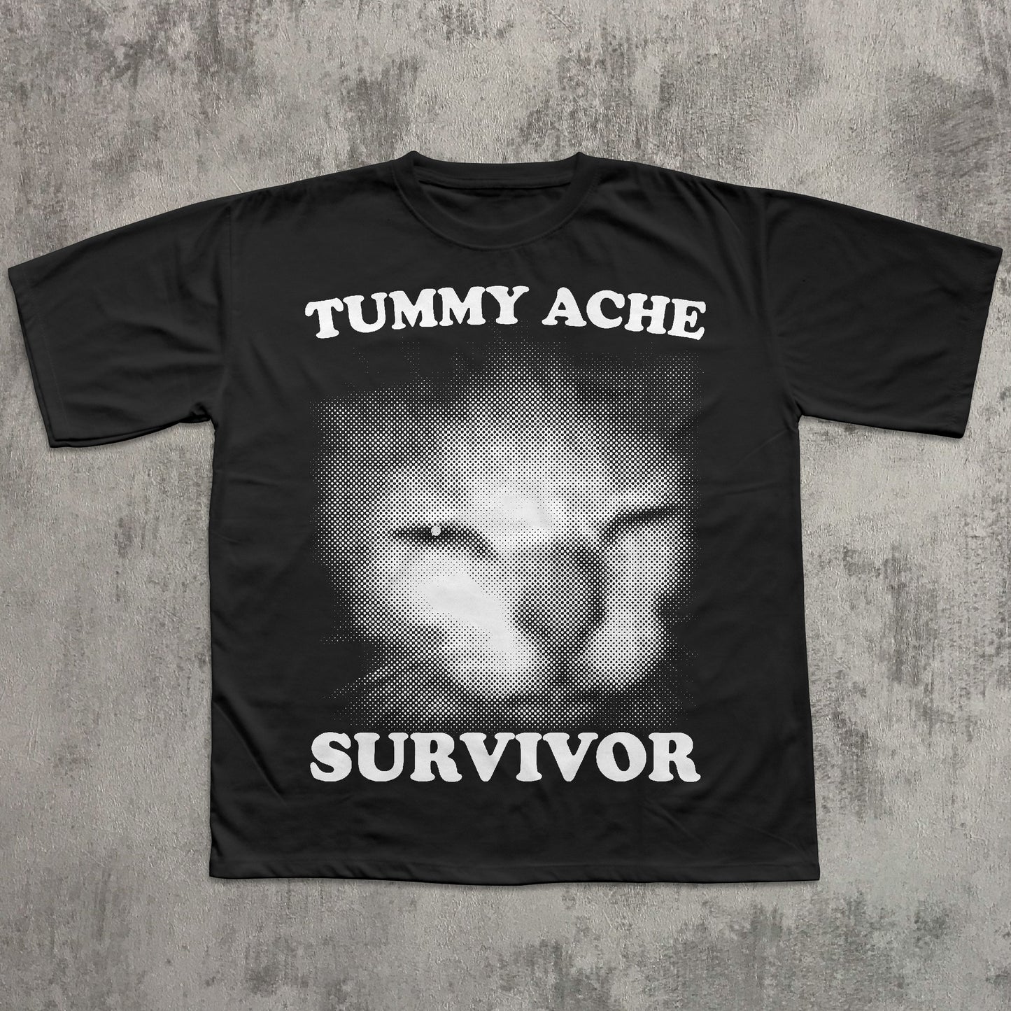 Black t-shirt with a graphic of a cat's face and text 'TUMMY ACHE SURVIVOR' on a textured gray background