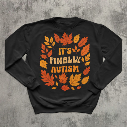 Black sweatshirt with autumn leaf design and 'It's Finally Autism' text on a textured gray background