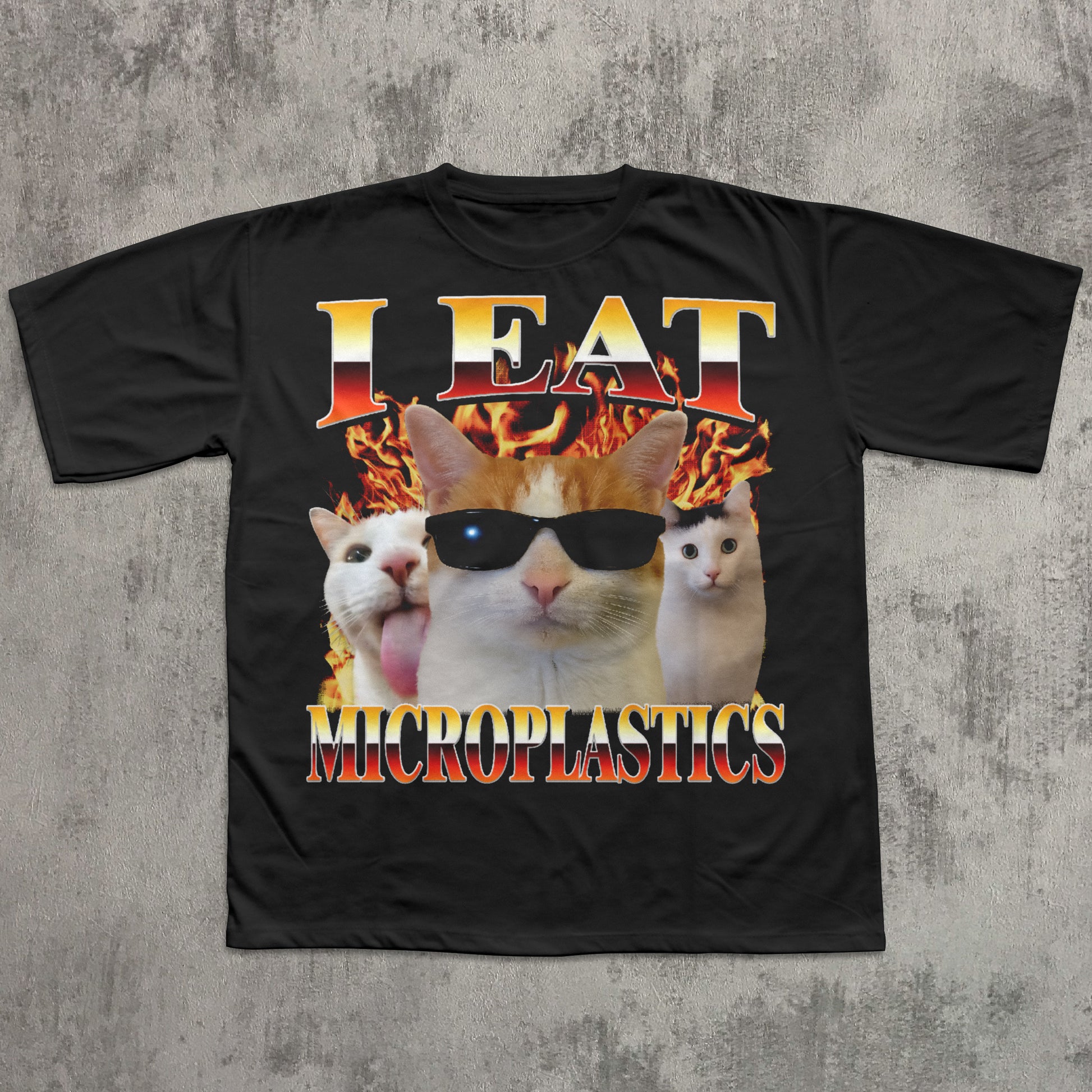 Black t-shirt with a graphic of cats wearing sunglasses and text 'I Eat Microplastics' on a concrete surface.