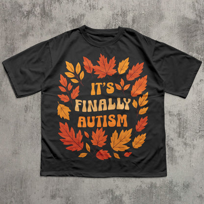 Black t-shirt with autumn leaf design and 'It's Finally Autism' text on a textured gray background