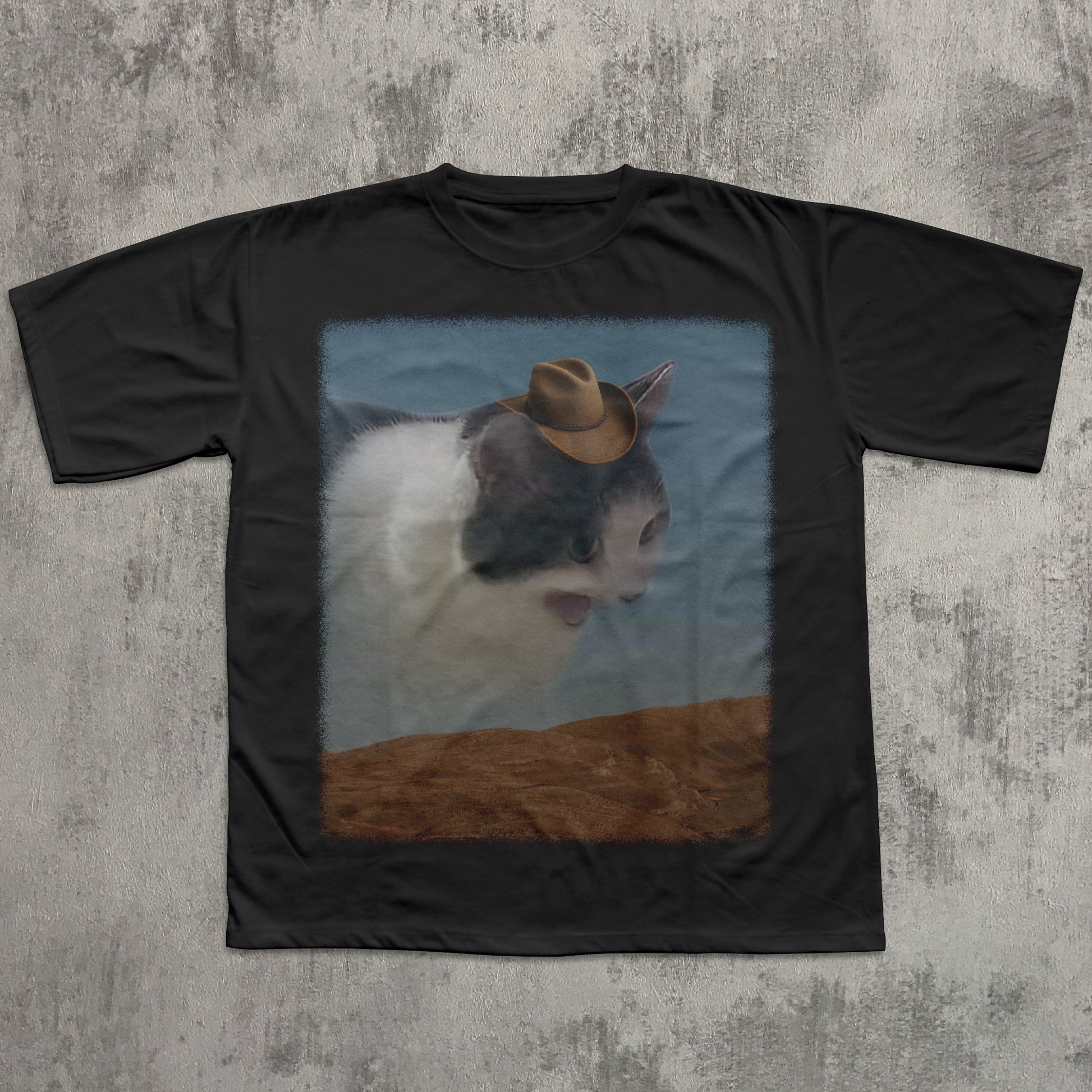 Black t-shirt with a cat wearing a cowboy hat graphic on a textured gray background