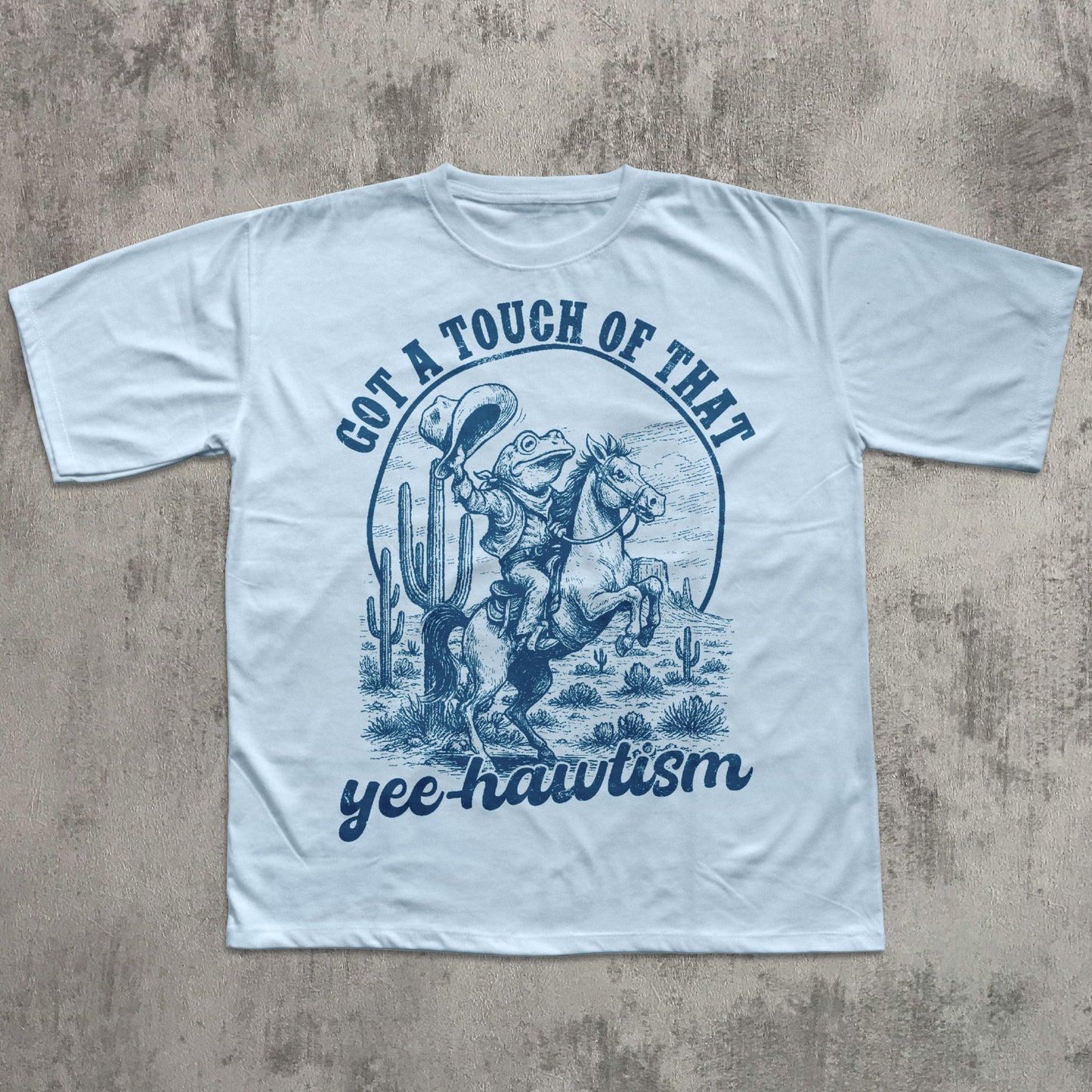 Blue T-shirt featuring a blue vintage-style illustration of a cowboy frog riding a rearing horse in a desert scene with cacti, with the text ‘Got a touch of that yee-hawtism’ above and below the graphic.
