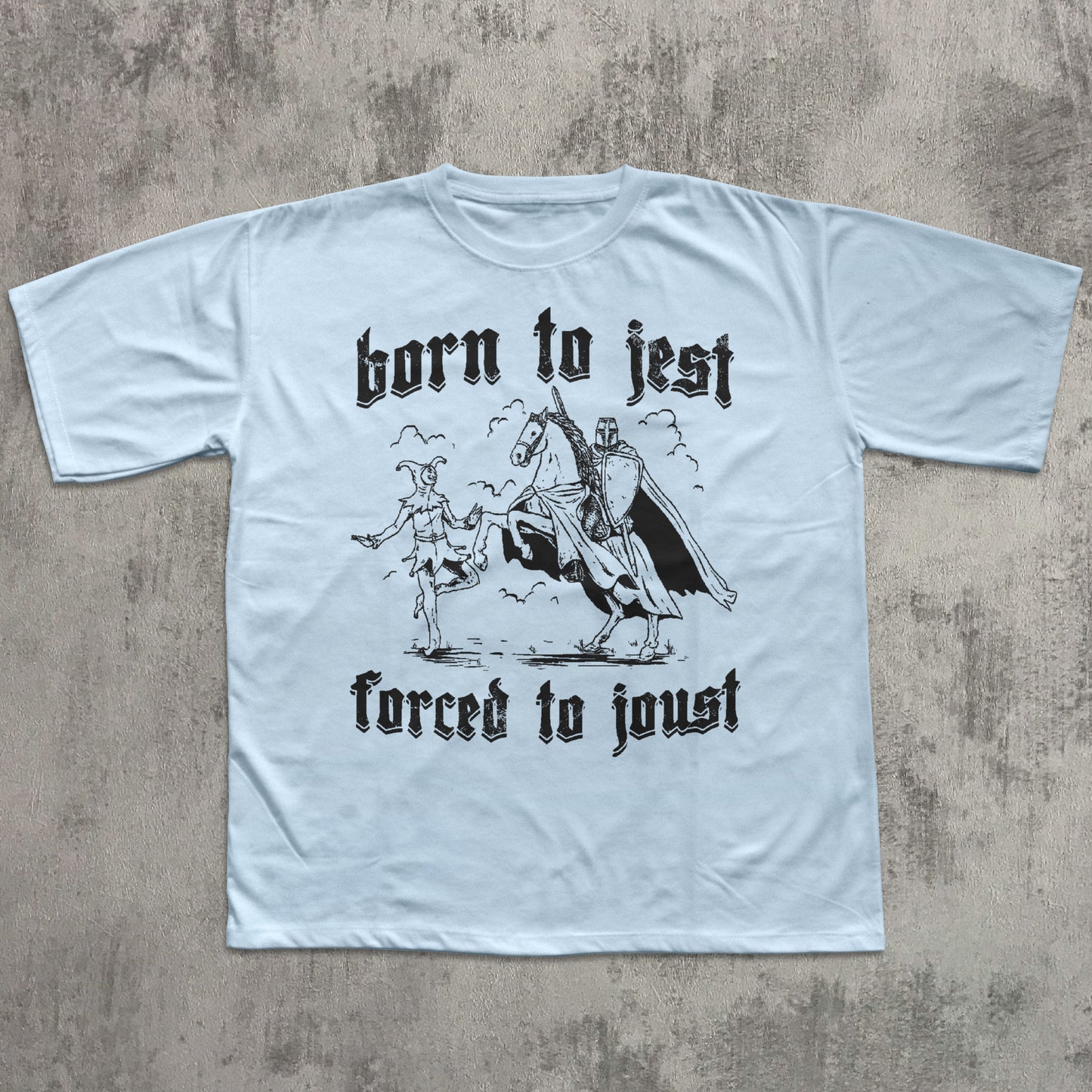 Light blue t-shirt with black graphic and text 'born to jest, forced to joust' on a textured gray background