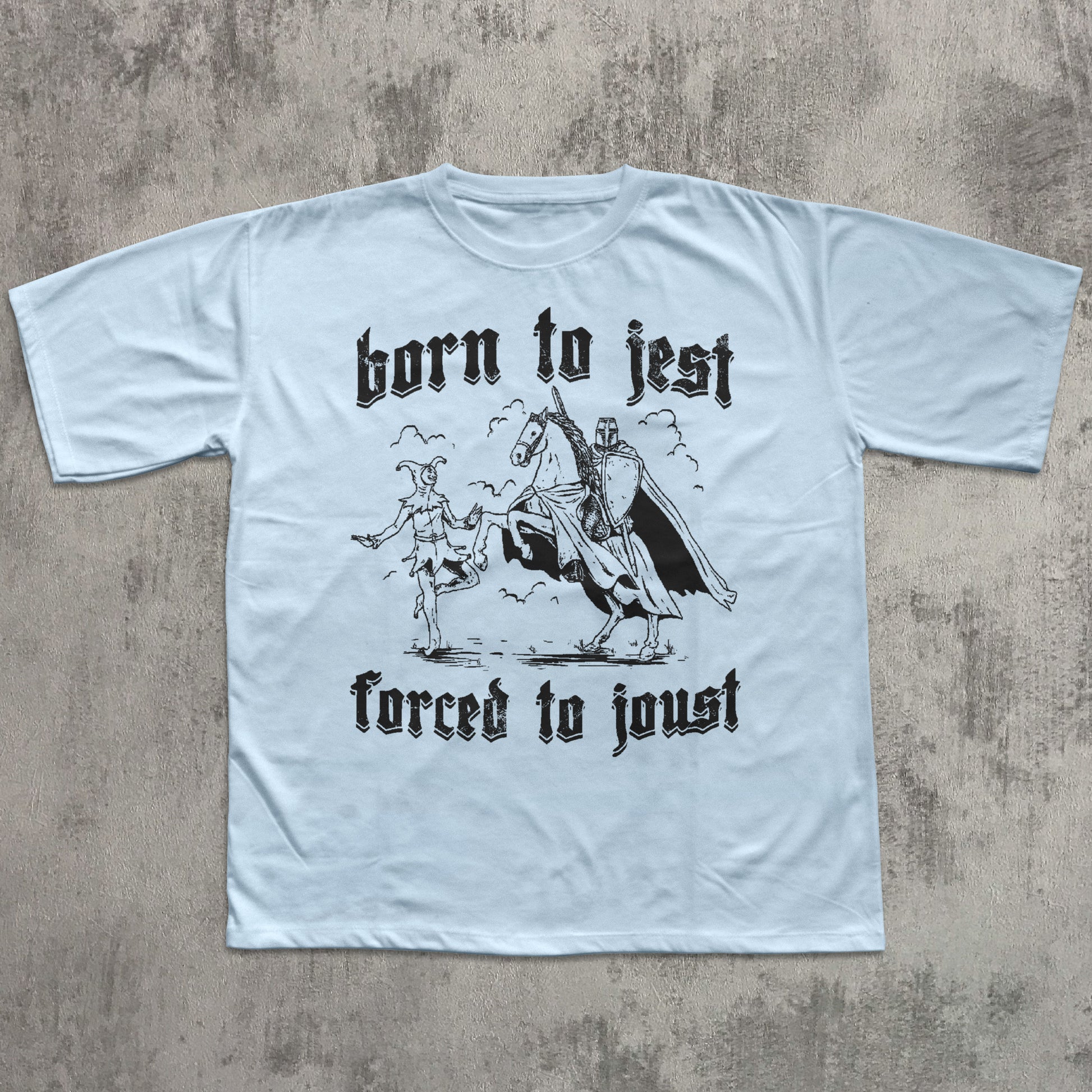 Light blue t-shirt with black graphic and text 'born to jest, forced to joust' on a textured gray background