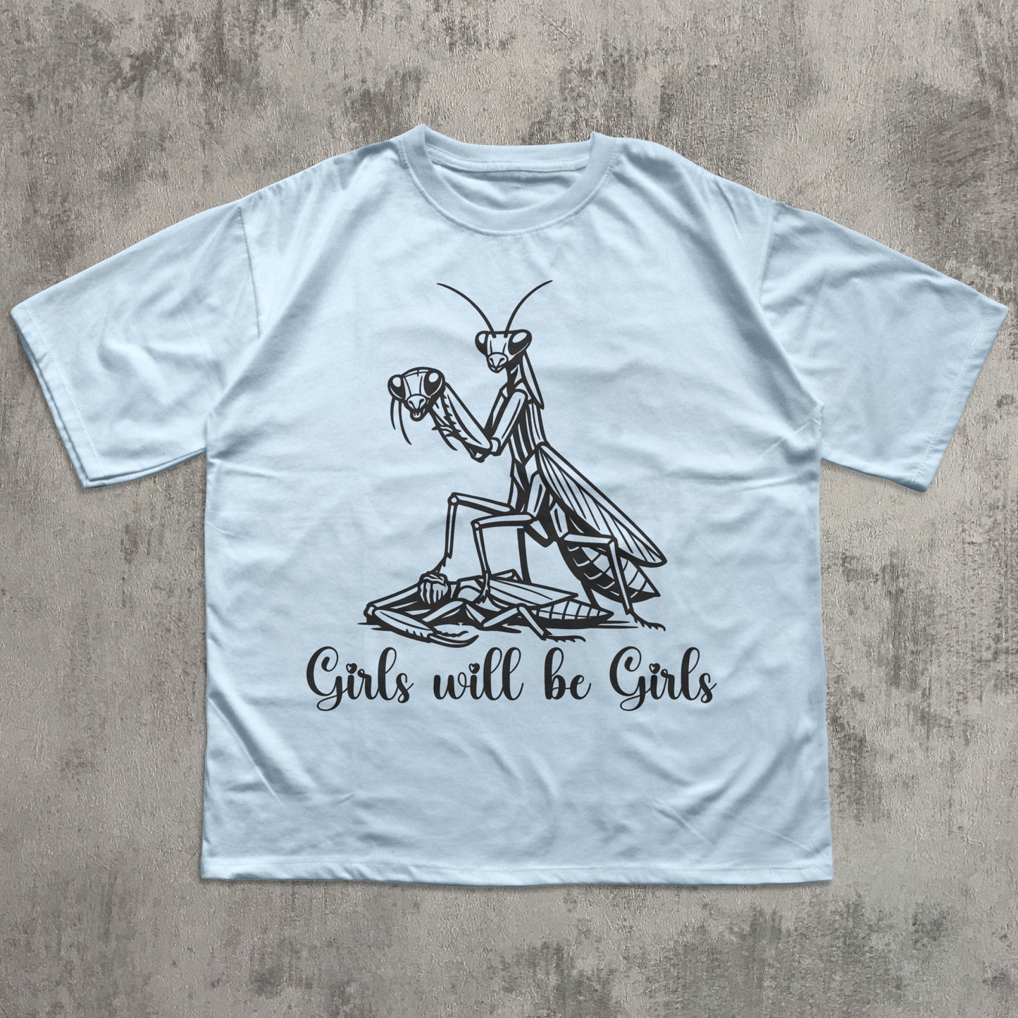 Light blue t-shirt with a praying mantis graphic and 'Girls will be Girls' text on a textured surface.