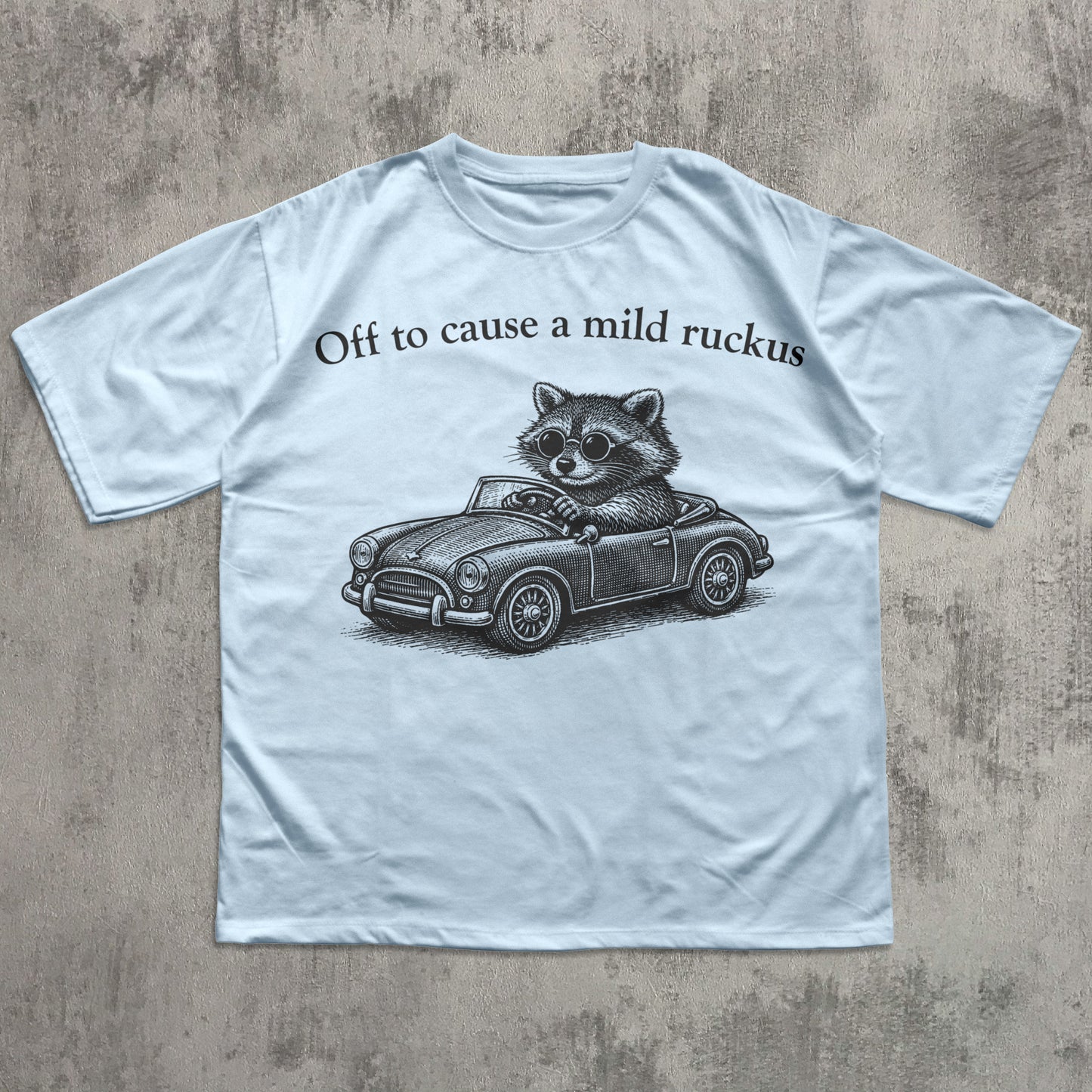 Light blue t-shirt with a raccoon driving a car graphic and text on a textured gray background