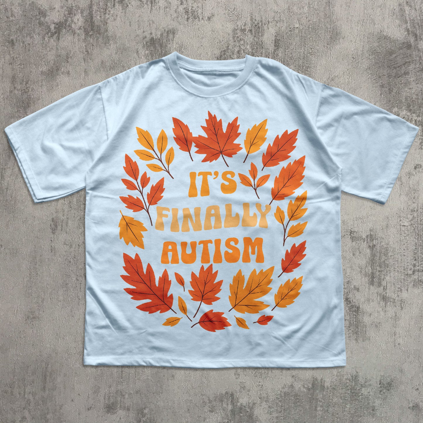 White t-shirt with autumn leaf design and 'It's Finally Autism' text on a concrete surface