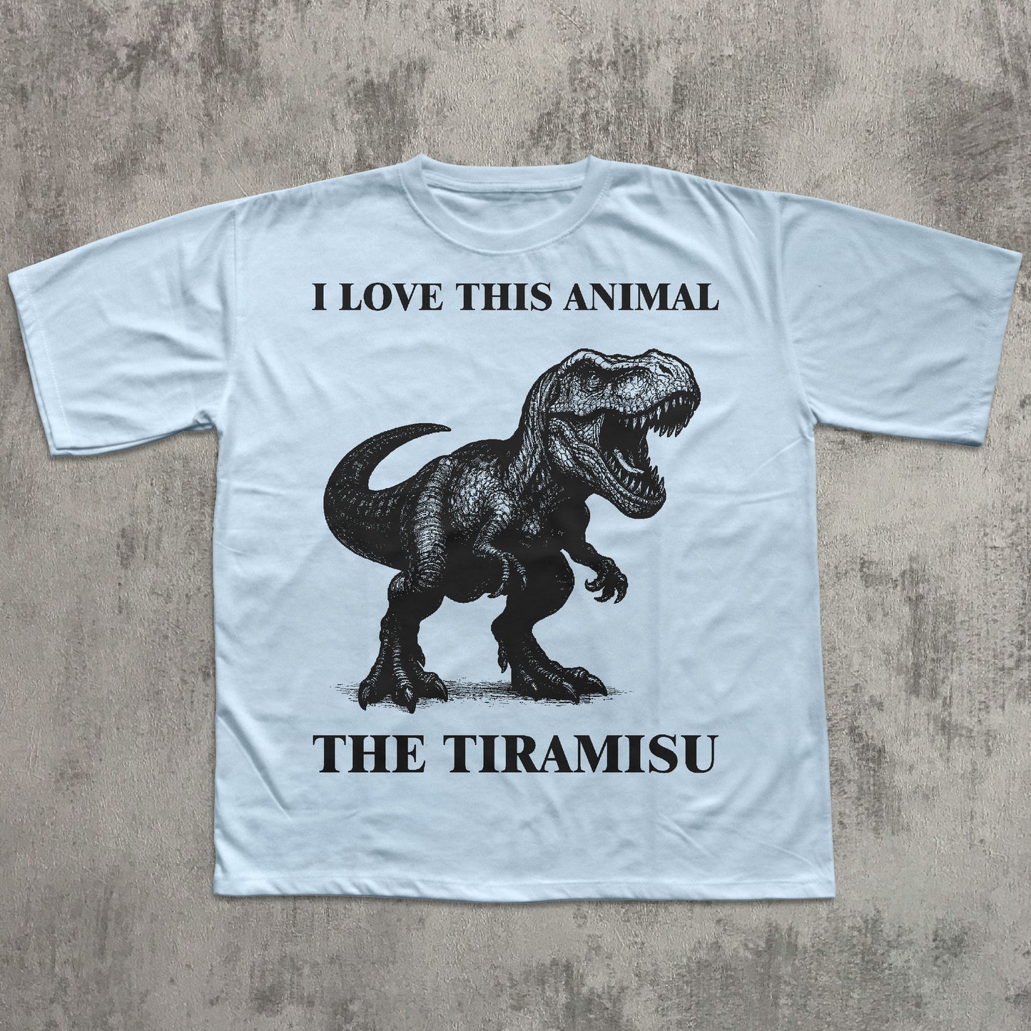 Light blue t-shirt with a dinosaur graphic and text on a concrete surface