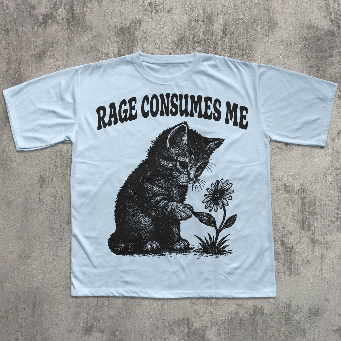 Light blue t-shirt with a graphic of a kitten holding a flower and text 'RAGE CONSUMES ME' on a concrete surface.
