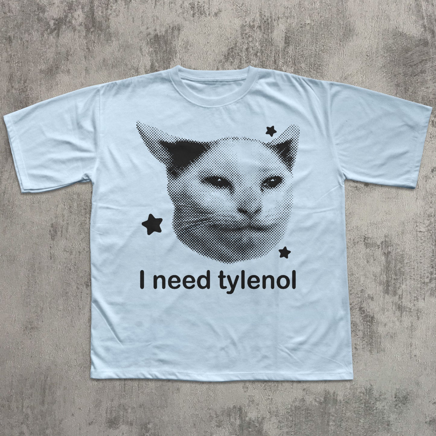 White t-shirt with a graphic of a cat's face and text 'I need tylenol' on a textured gray background
