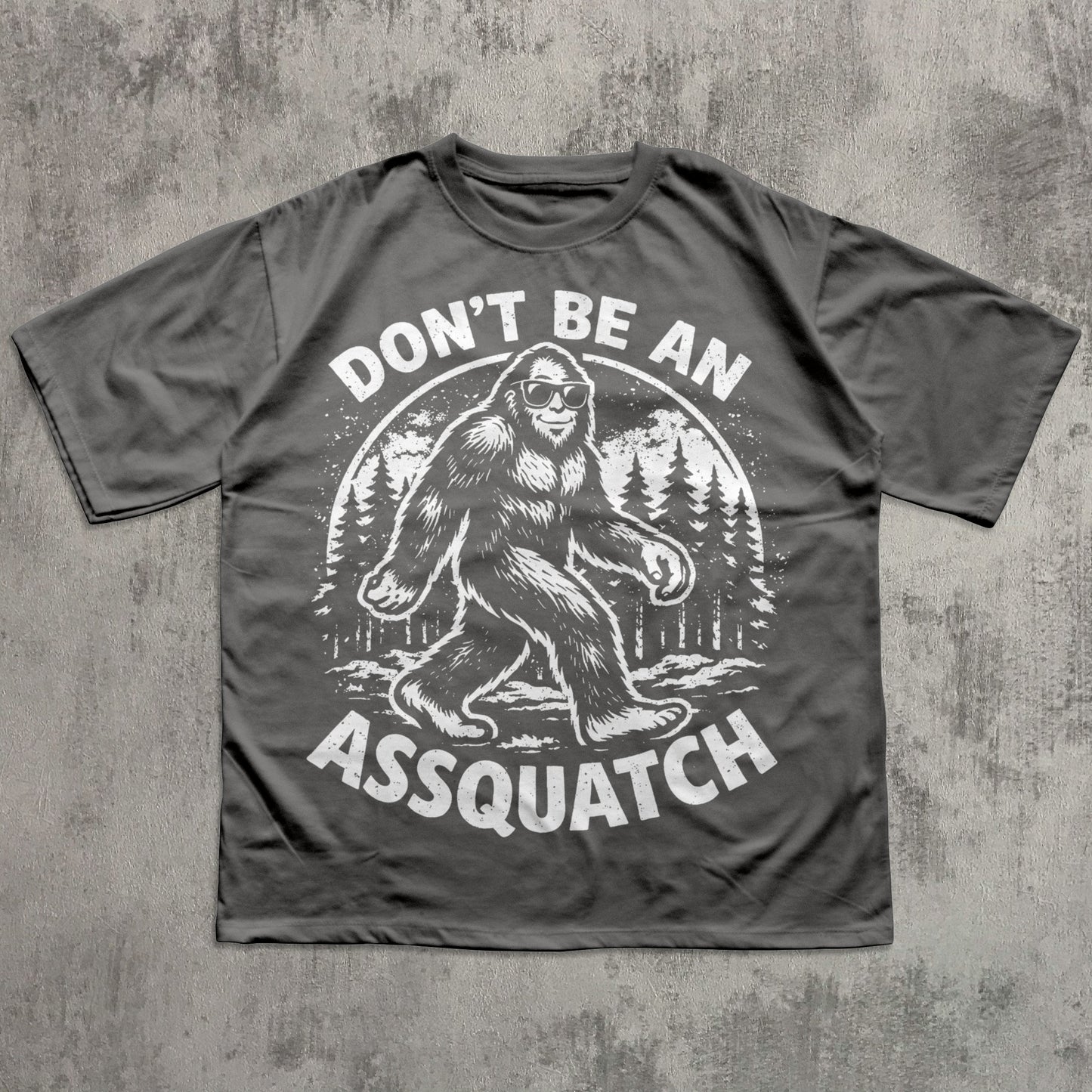 Gray t-shirt with a graphic of a Sasquatch and text 'Don't Be An Assquatch' on a concrete background