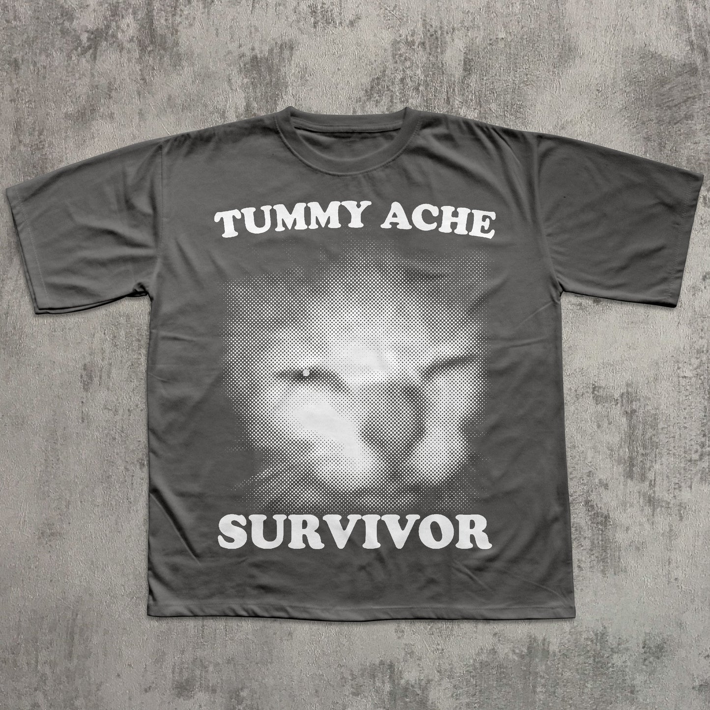 Gray t-shirt with a graphic of a cat and text 'TUMMY ACHE SURVIVOR' on a textured gray background