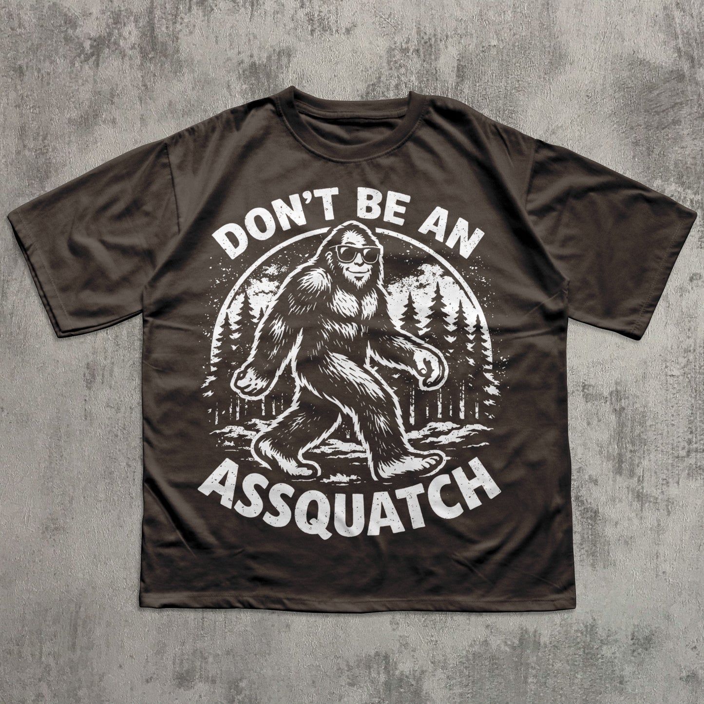 Brown t-shirt with a graphic of a bigfoot and text 'Don't Be An Assquatch' on a concrete surface.