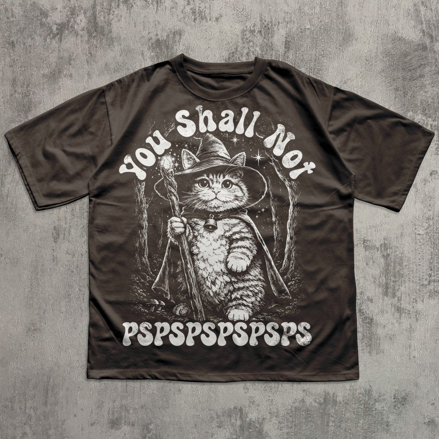 Brown t-shirt with a cat in a witch hat graphic and text on a textured gray background