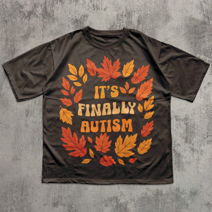 Brown t-shirt with autumn leaf design and 'It's Finally Autism' text on a gray textured background