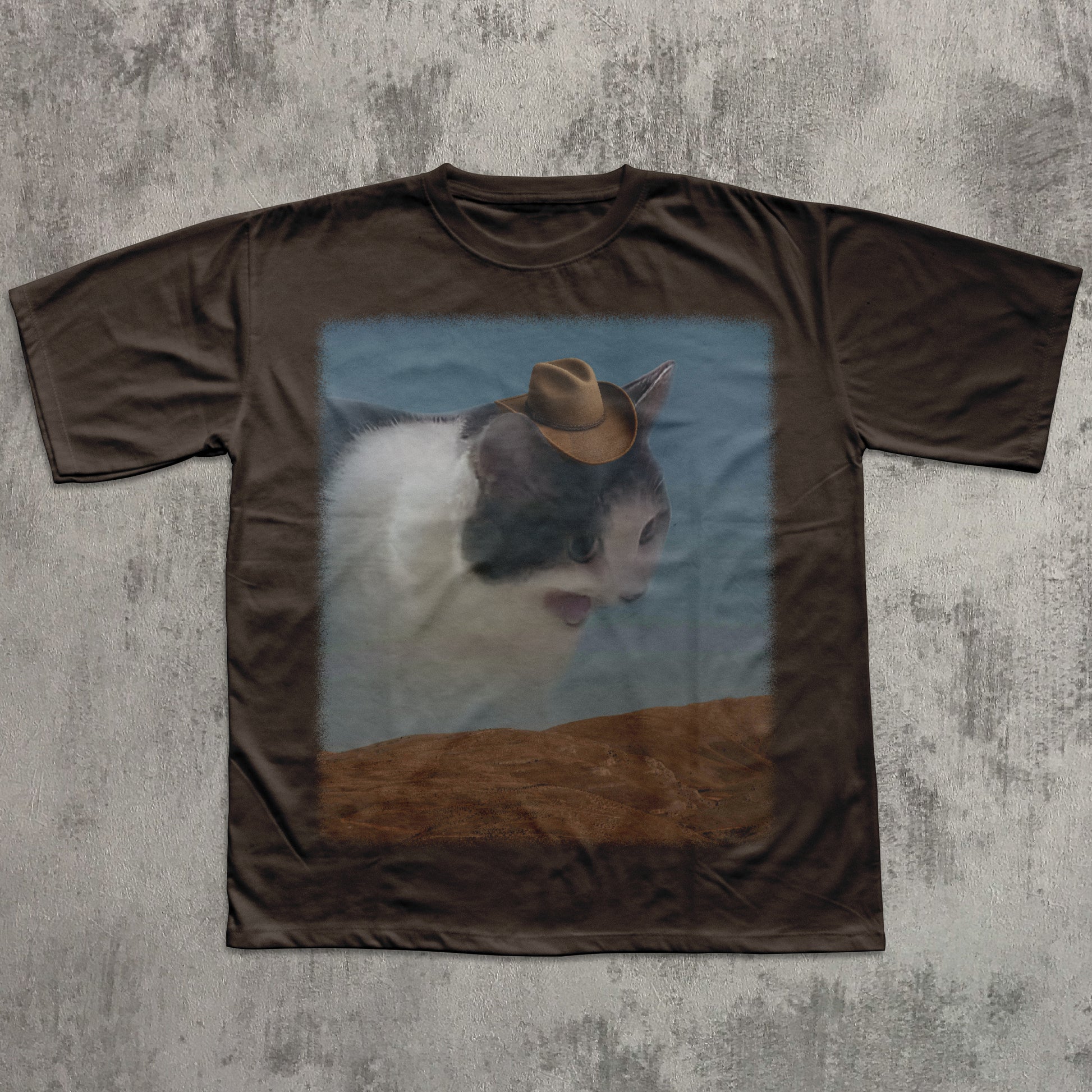 Brown t-shirt with a graphic of a cat wearing a cowboy hat on a textured gray background