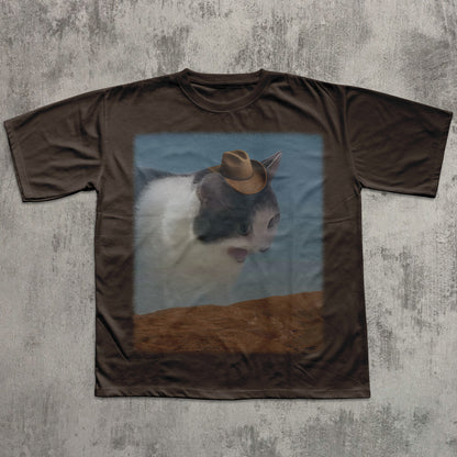 Brown t-shirt with a graphic of a cat wearing a cowboy hat on a textured gray background