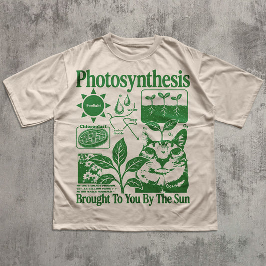 A beige short-sleeve t-shirt with a green graphic print titled 'Photosynthesis' featuring a cat illustration and educational diagrams, laid flat on a textured gray background.