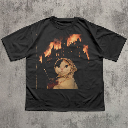 Black t-shirt with a graphic of a cat in a hood and a castle on fire, on a textured gray background.
