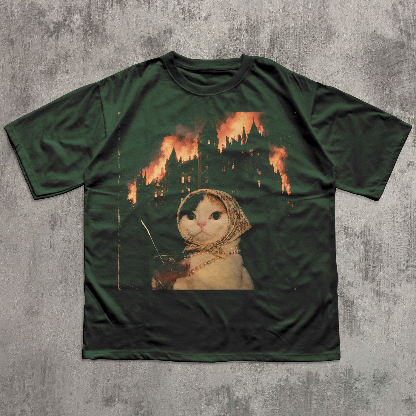 Green t-shirt with a graphic of a cat in a hood and a castle on fire, on a textured gray background.