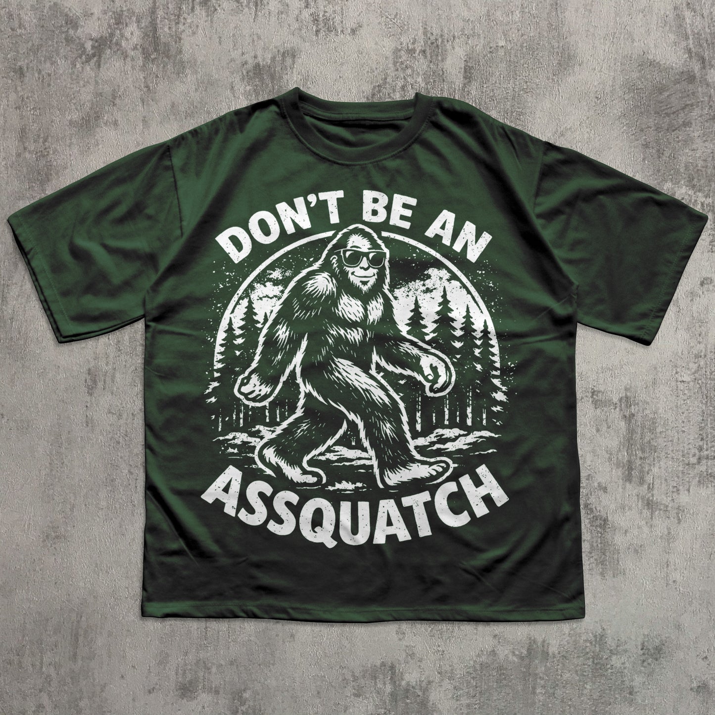 Green t-shirt with a graphic of a Sasquatch and text 'Don't be an Assquatch' on a concrete surface.