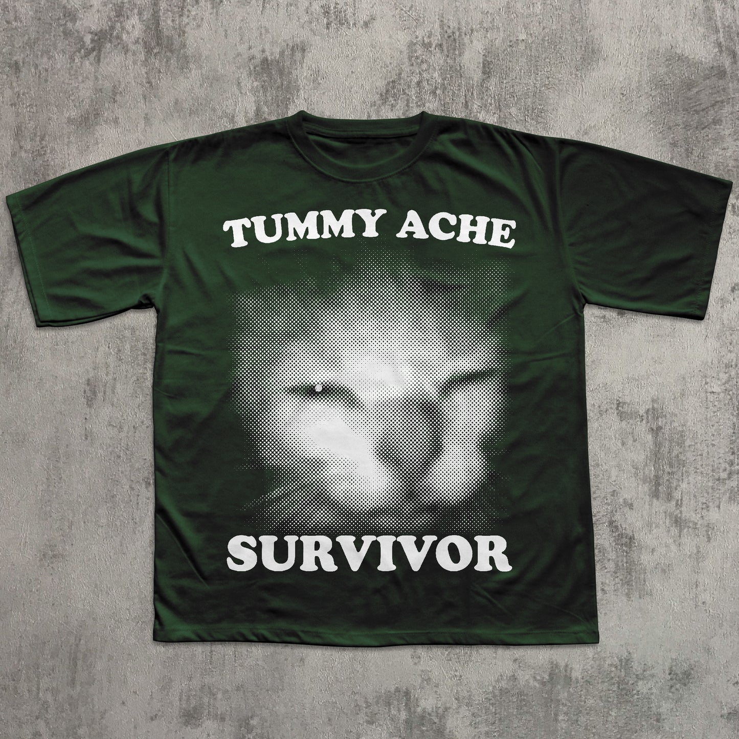 Green t-shirt with a cat face graphic and text 'TUMMY ACHE SURVIVOR' on a textured gray background