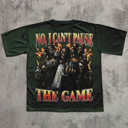 Green t-shirt with graphic design of skeletons holding guns and a pause button, text 'No, I Can't Pause The Game' on a textured gray background.