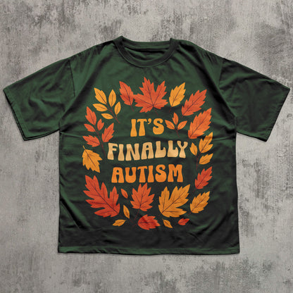 Green t-shirt with autumn leaf design and 'It's Finally Autism' text on a textured gray background