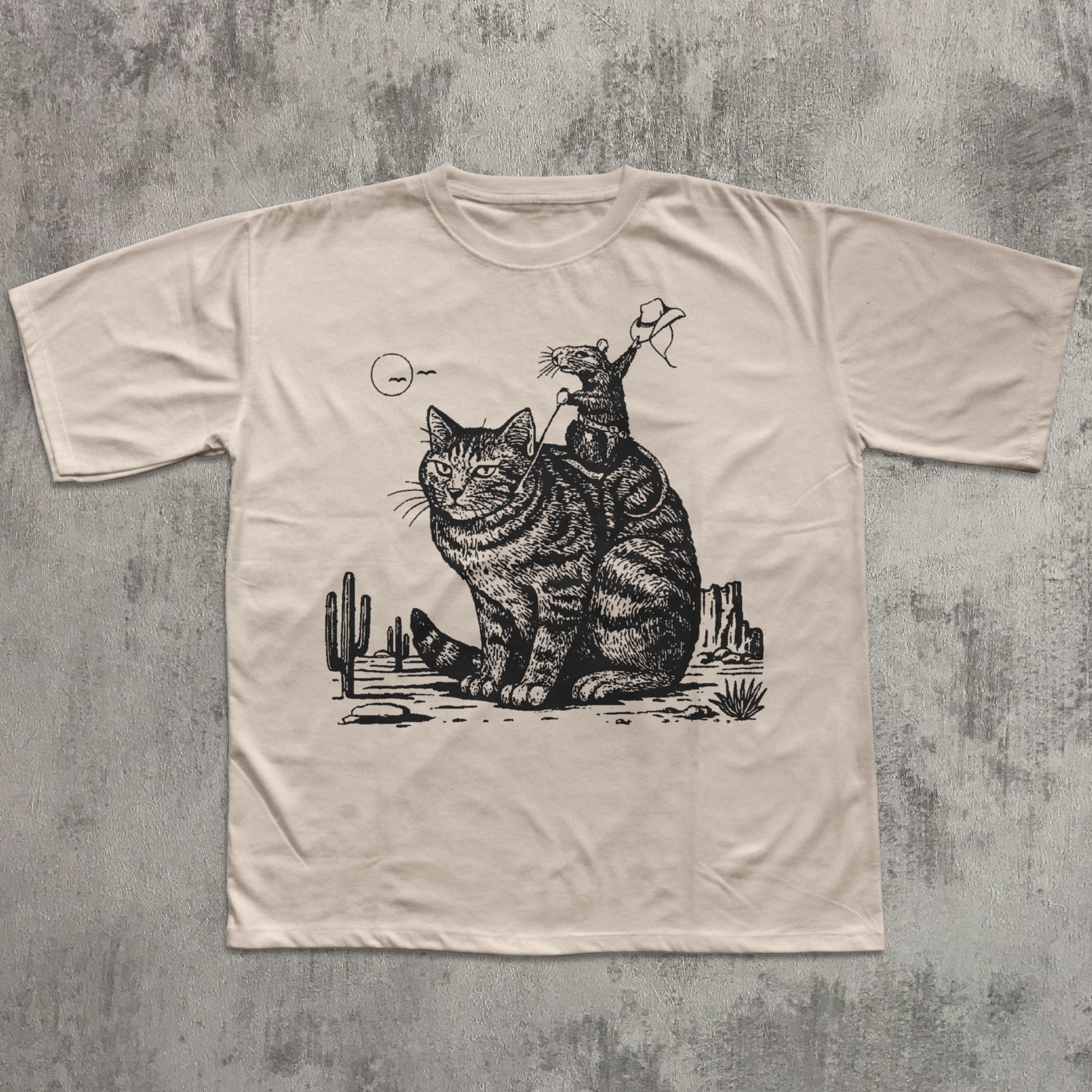 Beige t-shirt with a graphic of two cats and cacti on a textured gray background