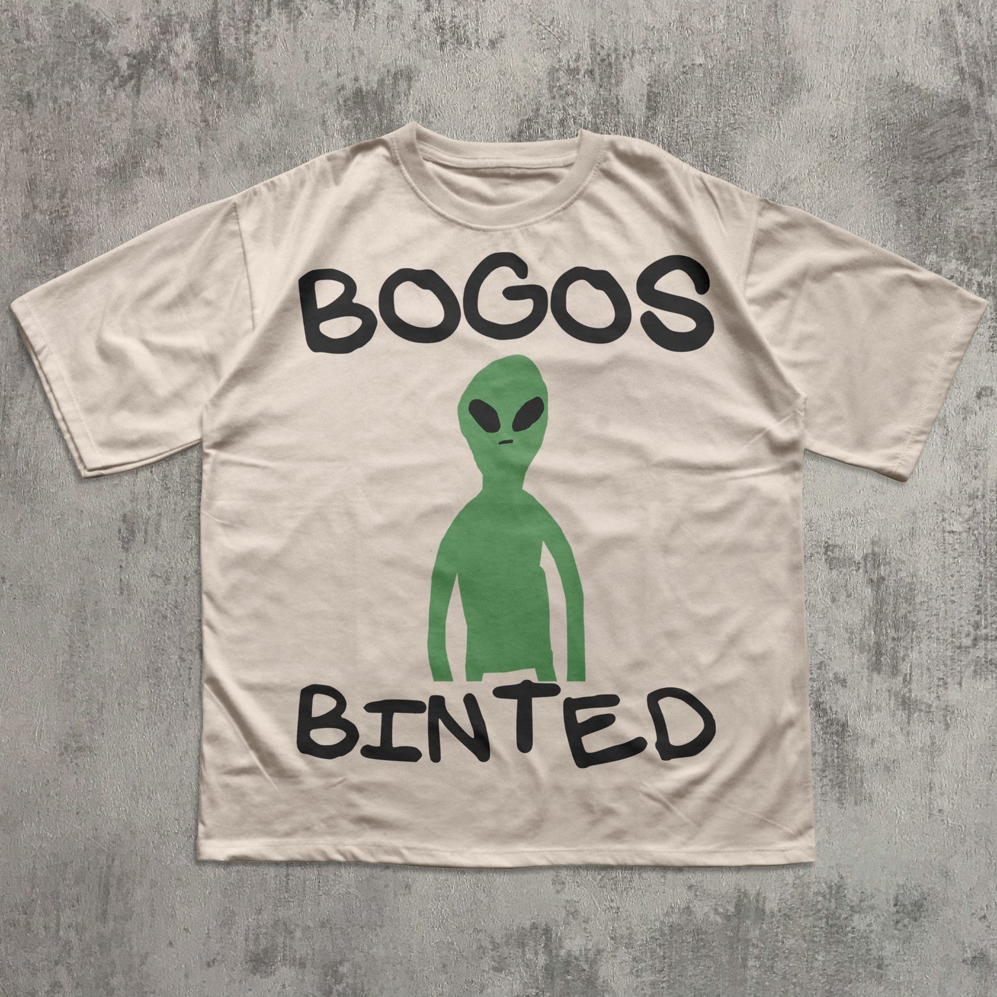 Beige t-shirt with a green alien graphic and text 'BOGOS BINTED' on a textured gray background