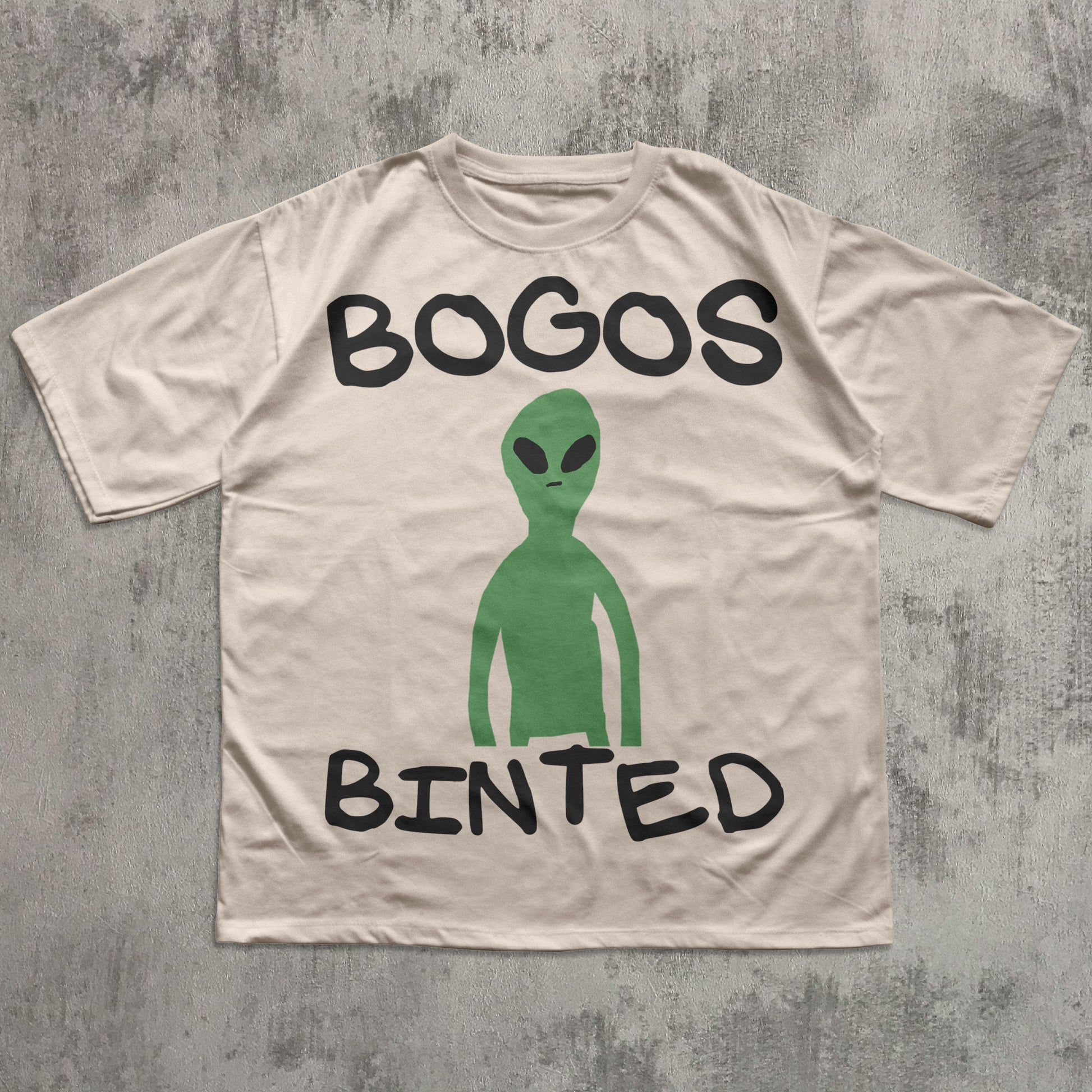Beige t-shirt with a green alien graphic and text 'BOGOS BINTED' on a textured gray background
